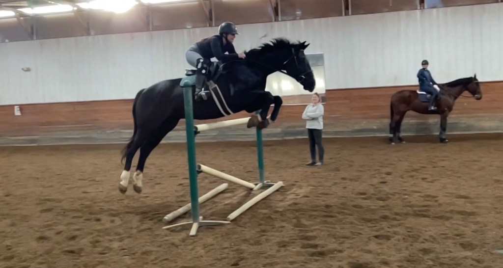 Caroline Wollman riding her horse, Cody, in an indoor equestrian arena. The rider is on a black horse that's jumping over an obstacle with green poles and wooden beams. A person observes behind them and a second rider on a brown horse watches from the background.