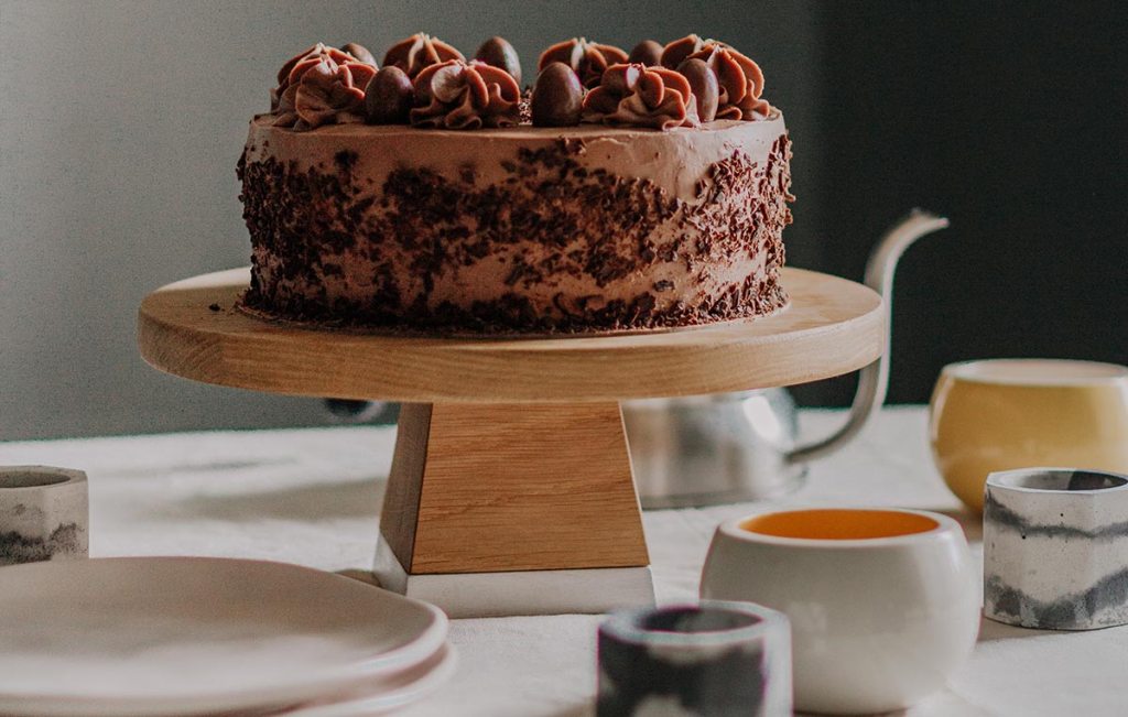 A chocolate cake sitting on a wooden cake stand with various cups and dishes surrounding. 