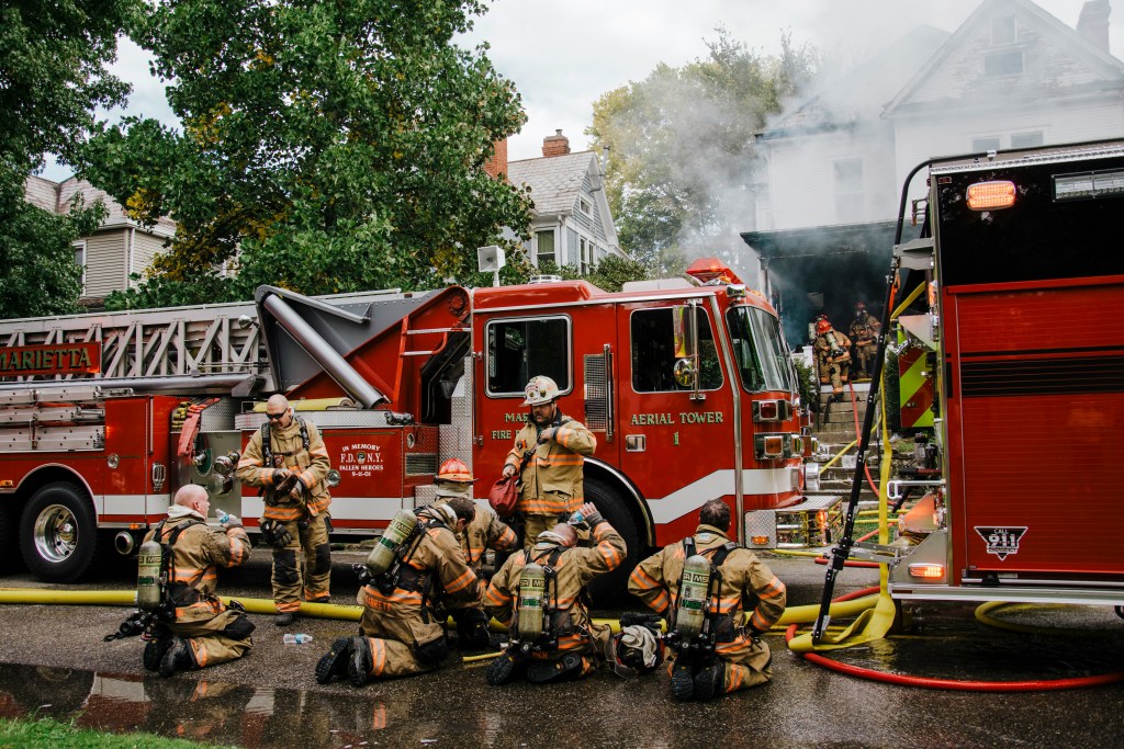 Firefighters in full gear rest near red fire trucks as smoke pours from a white house behind them, while other crew members continue working at the scene.