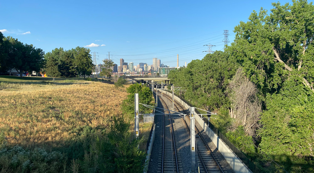 View of the Denver city skyline in the distance with a railroad surrounded by greenery in the foreground.