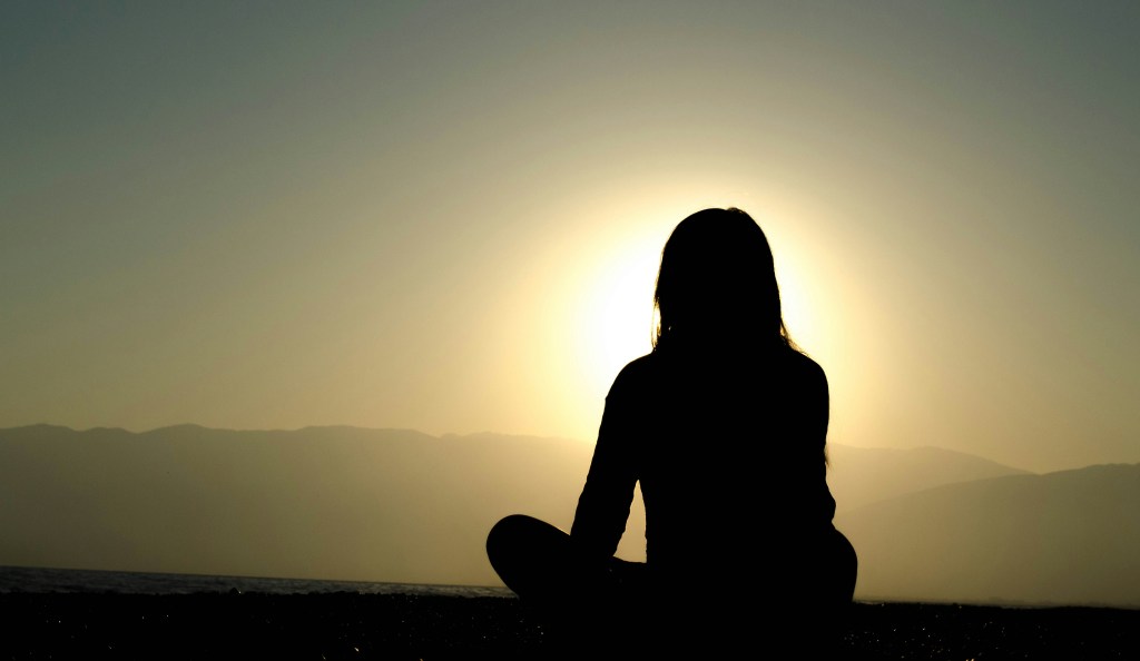 Silhouette of a person meditating outdoors at sunrise or sunset, with mountains and water in the background.