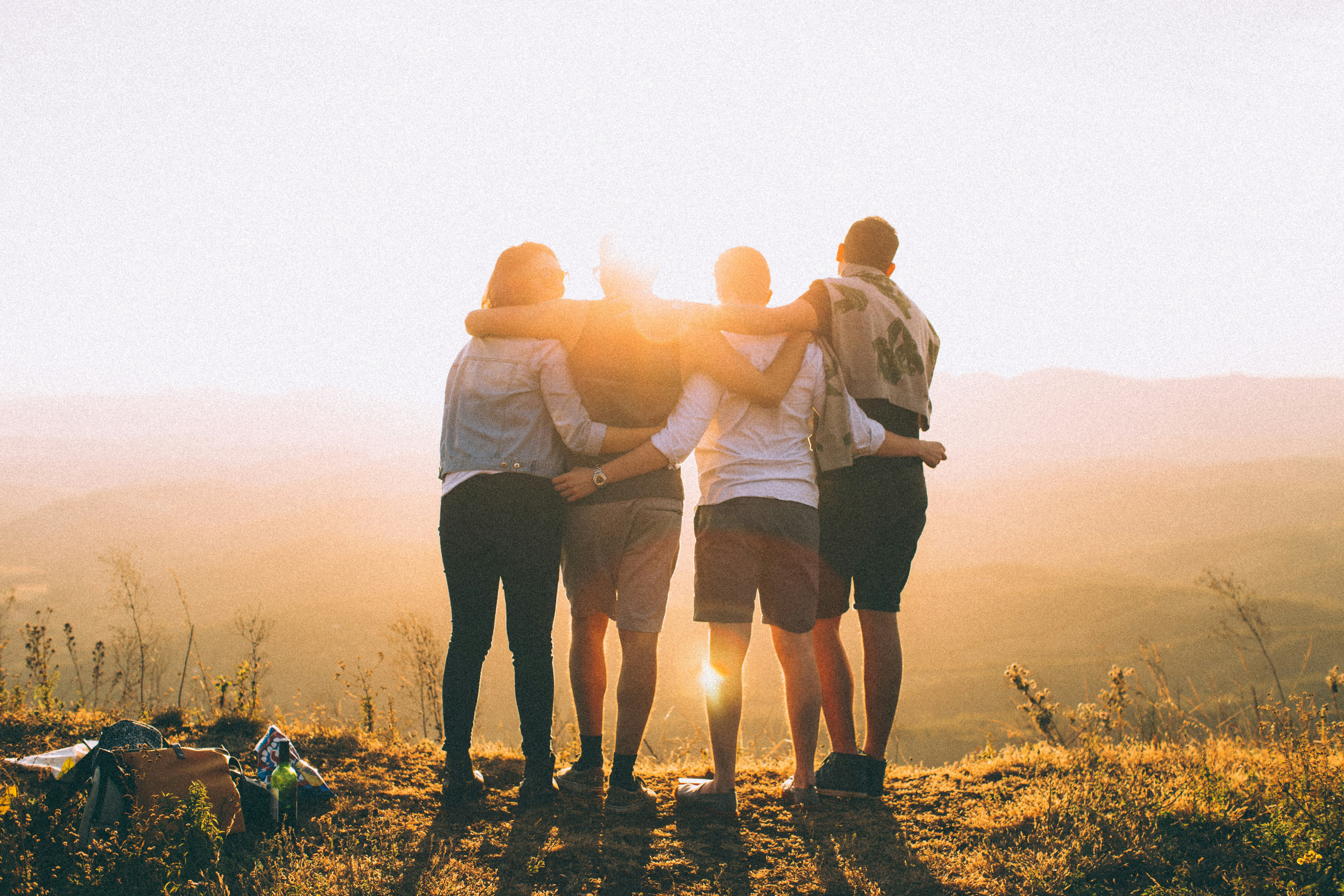 Four friends stand on a hilltop with their arms around each other, looking toward the sunset over a scenic mountain landscape.