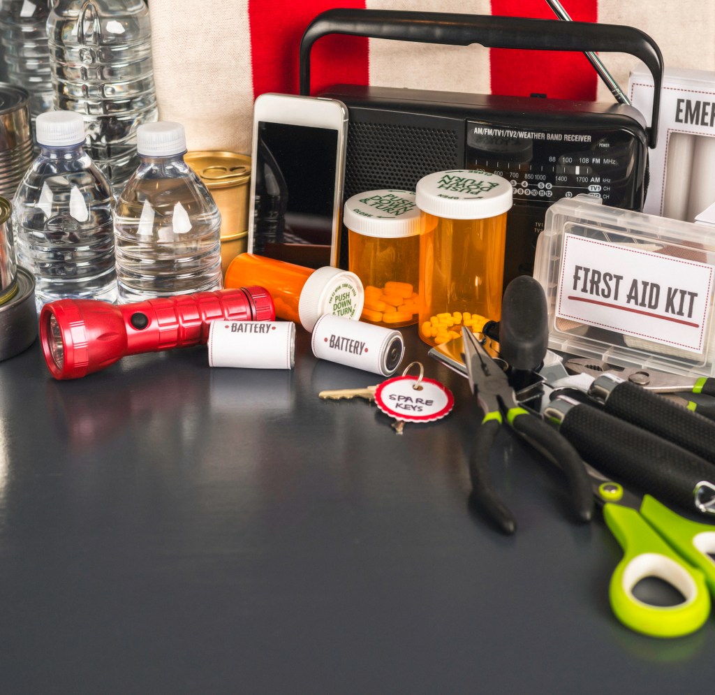 Close-up of emergency preparedness supplies on a table, including water bottles, medication, flashlight, batteries, tools, a first aid kit, spare keys, and a radio.