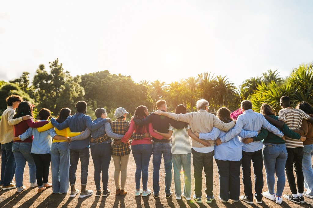 A diverse group of people standing side by side with arms around each other, facing the sun in a lush outdoor setting.