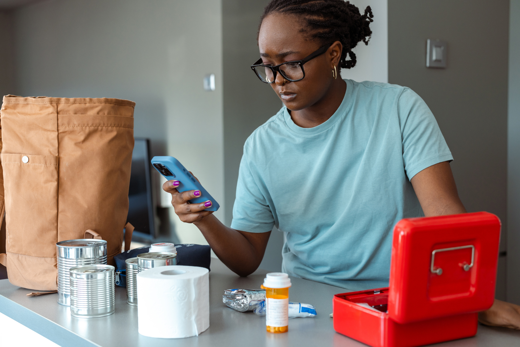 Person looking at their phone while organizing emergency supplies, including canned food, medicine, and a red first aid kit on a counter.