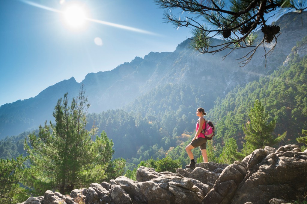 A woman wearing hiking gear and a pink backpack stands confidently on a rocky outcrop, overlooking a lush, green mountain landscape.