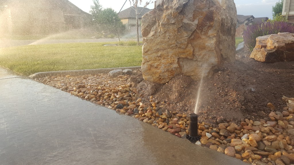 An active sprinkler being blocked by a large rock disrupting the path the water should have. Sprinklers in the background are watering a lawn properly with no blockages.