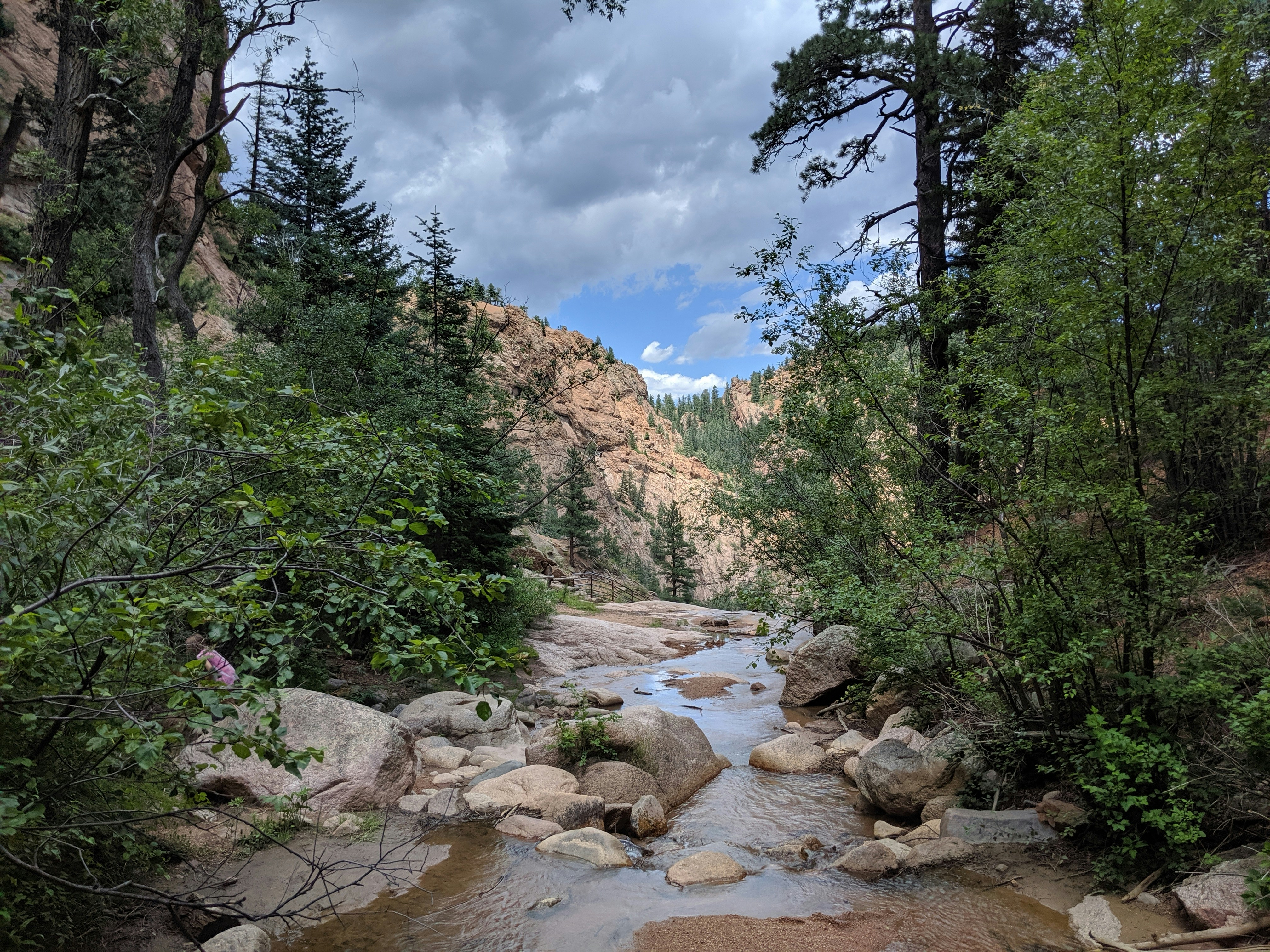A shallow mountain stream flows over smooth rocks through a forested canyon, with pine trees and dense green foliage on either side. Towering rocky cliffs rise in the background under a dramatic cloudy sky with a patch of blue peeking through.