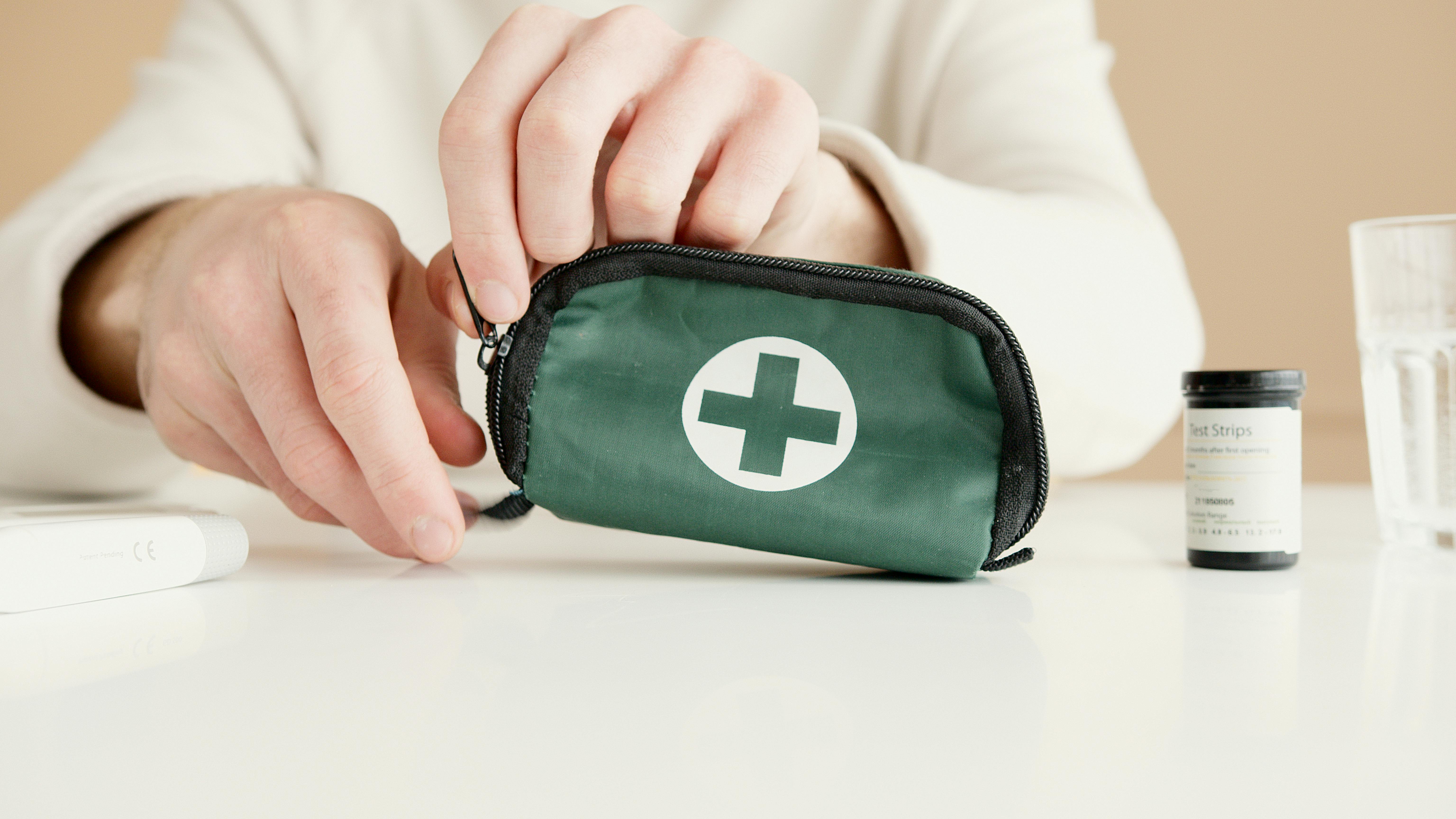 Close-up of a person’s hand opening a small green first aid kit with a white cross symbol on it, placed on a white surface next to a glass of water and a container of test strips.