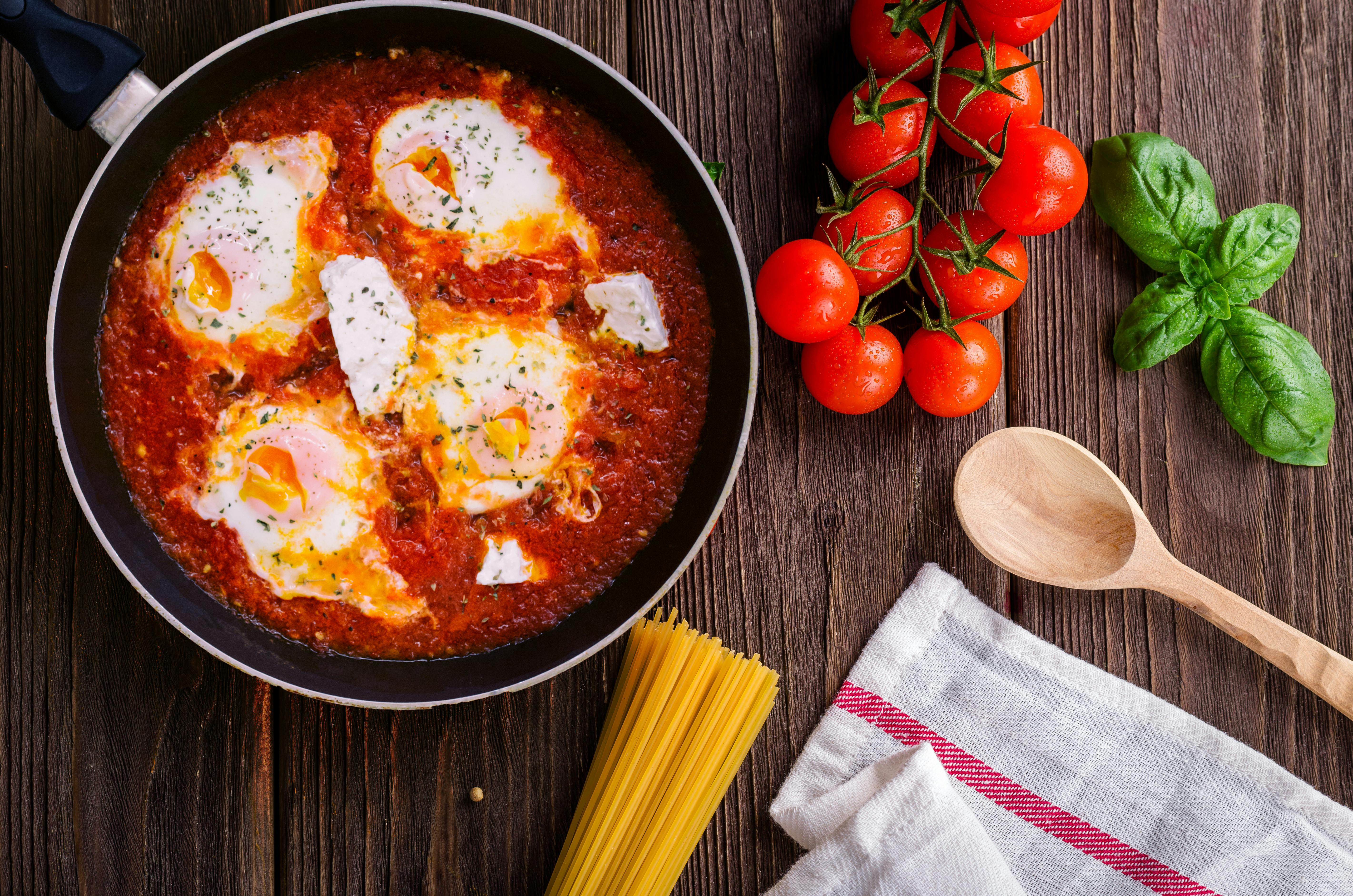 Pan of shakshuka with poached eggs in tomato sauce, surrounded by cherry tomatoes, fresh basil, and spaghetti on a rustic wooden table.