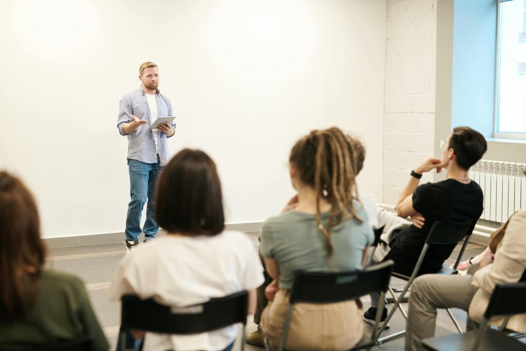 A man stands in front of a small seated audience, holding a tablet and speaking during a presentation or workshop in a modern, well-lit room.