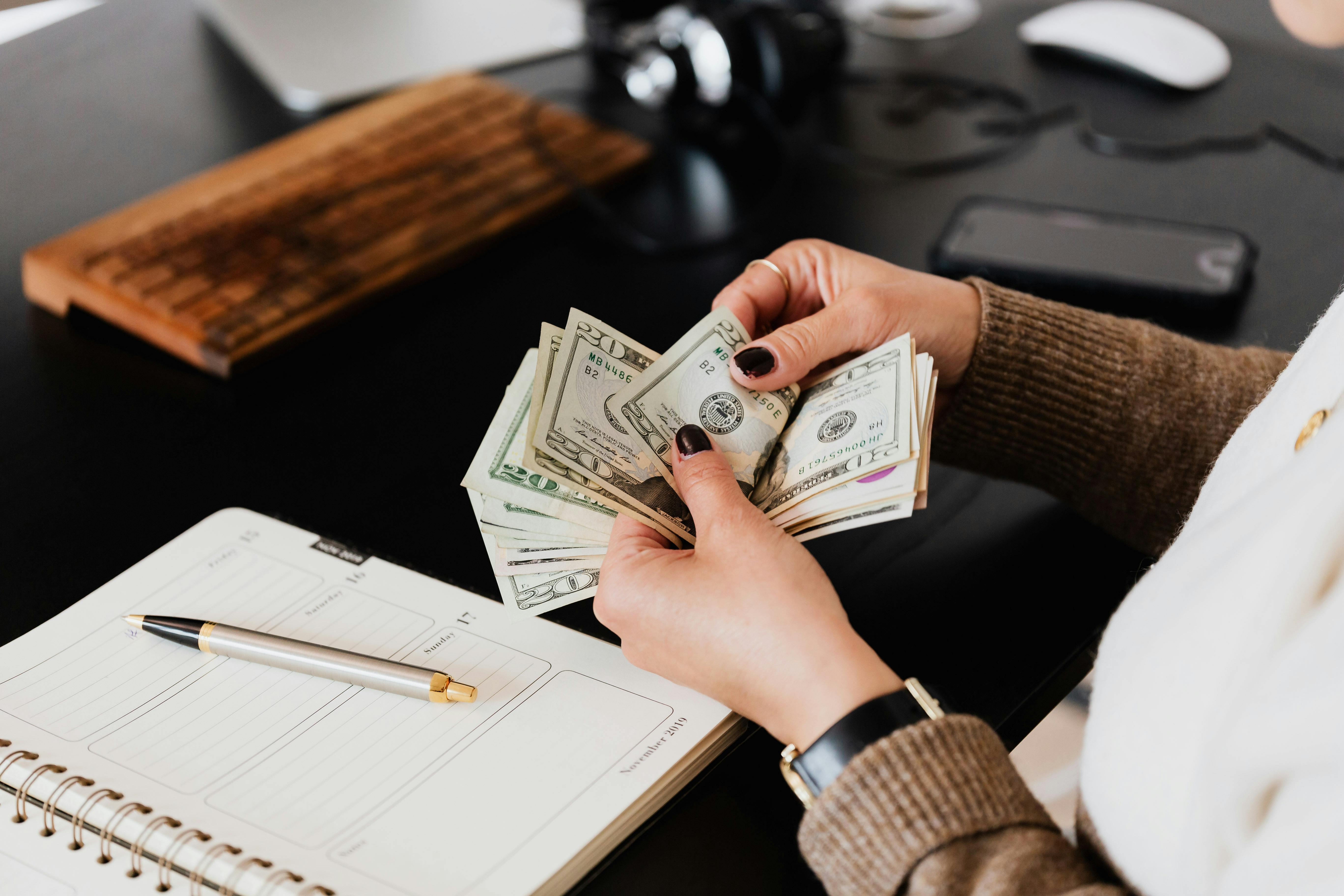 Person counting a stack of US dollar bills at a black desk with a spiral planner, pen, and wooden keyboard nearby.