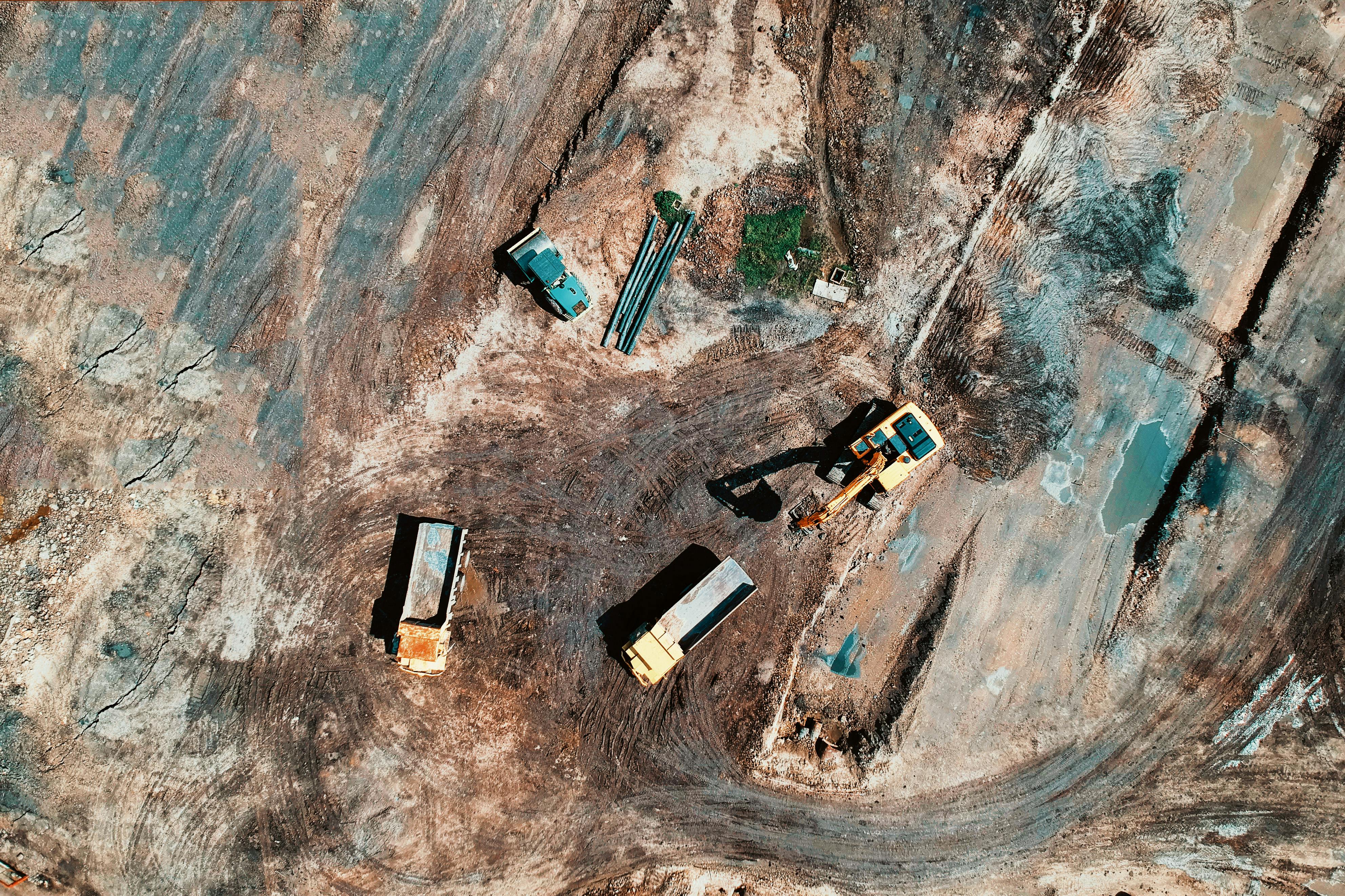 An aerial view of a mining site showing earth-moving equipment, including two trucks and an excavator, on a dirt-covered landscape with visible tire tracks and scattered materials.