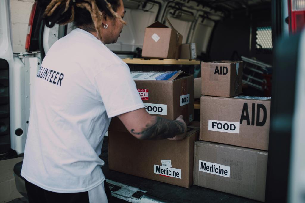 Volunteer loading cardboard boxes labeled “FOOD,” “AID,” and “Medicine” into the back of a delivery van.