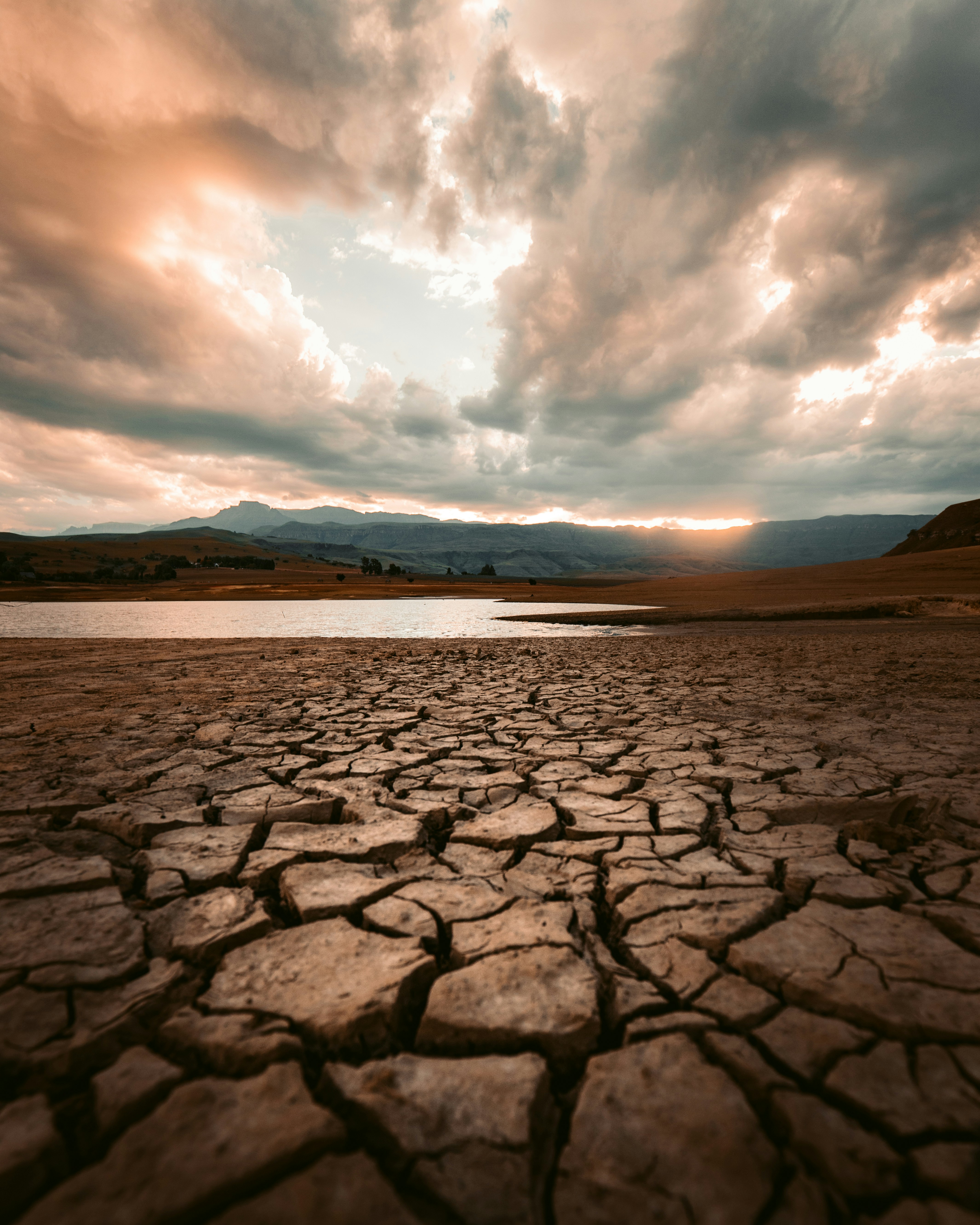 Cracked, parched earth stretches toward a shimmering body of water under a dramatic sky with thick, golden-lit clouds and distant mountain silhouettes at sunset.