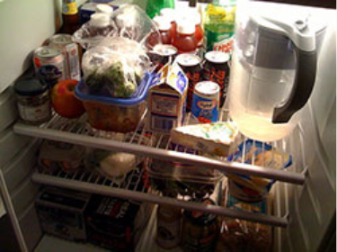 Open refrigerator with various drinks, condiments, produce, leftovers, and a water pitcher stored on wire shelves.