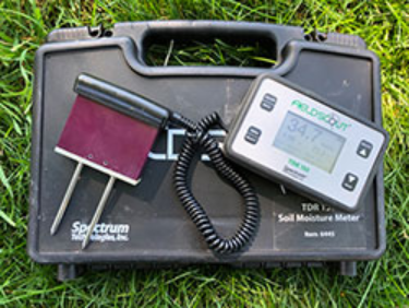 A portable soil moisture meter with probes, placed on top of its black carrying case on the grass.