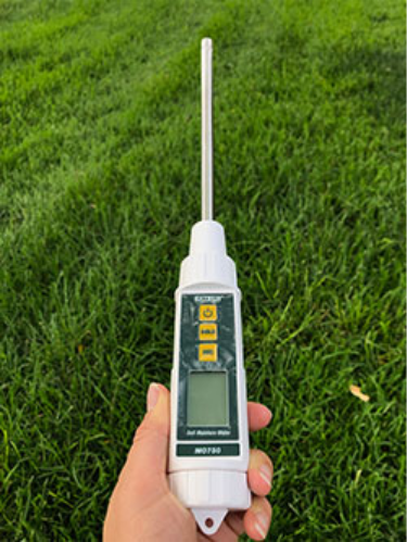A hand holding a digital soil moisture probe with a metal rod inserted into a green grassy area.