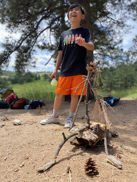 Smiling child outdoors in a forest, standing next to a small sculpture made of sticks, pinecones, and natural materials arranged on the ground.