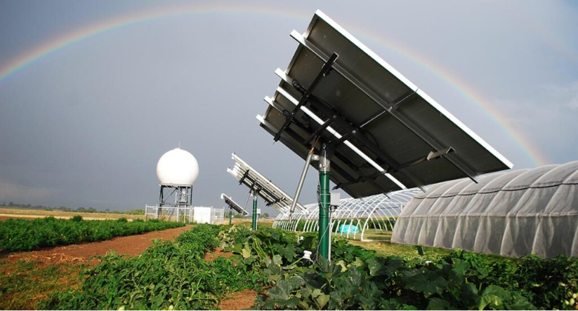 Field with solar panel rows and green crops beneath them, a white dome-shaped structure, greenhouses, and a rainbow in the sky.