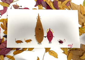 Art project where different colored leaves are arranged to look like a family of characters with faces and arms, placed on white paper surrounded by other scattered leaves.
