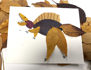 Creative art project showing a dinosaur made from pressed leaves glued to paper, with hand-drawn details like eyes, teeth, and legs.