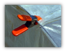 A photograph of a metal "duckbill" style clip with red handles and red grips on the clip end holding clear plastic sheeting onto a supportive wire frame.