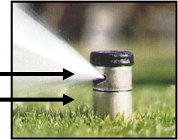 A photograph of a large rotor type irrigation head that is emitting water above a mown, green lawn. A black arrow indicates the point on the irrigation head where water is released; a second black arrow marks the top of the blades of grass; at least an inch is between the top of the lawn and the point of water emission.