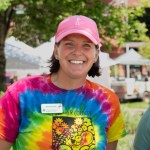 Alison O'Connor smiles at the camera wearing a pink hat and tie dye Larimer County Farmers Market t-shirt, standing in front of tents at the market in Fort Collins