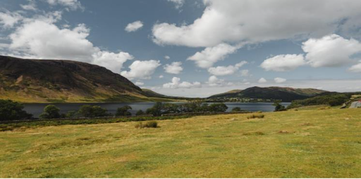 Large grassy field leading to a lake framed by rolling hills in the background. The sky is blue and partially cloudy.