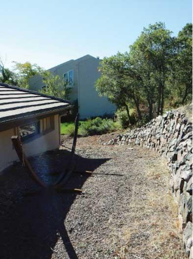 Side of a house with a rock wall and trees lining the right side. 