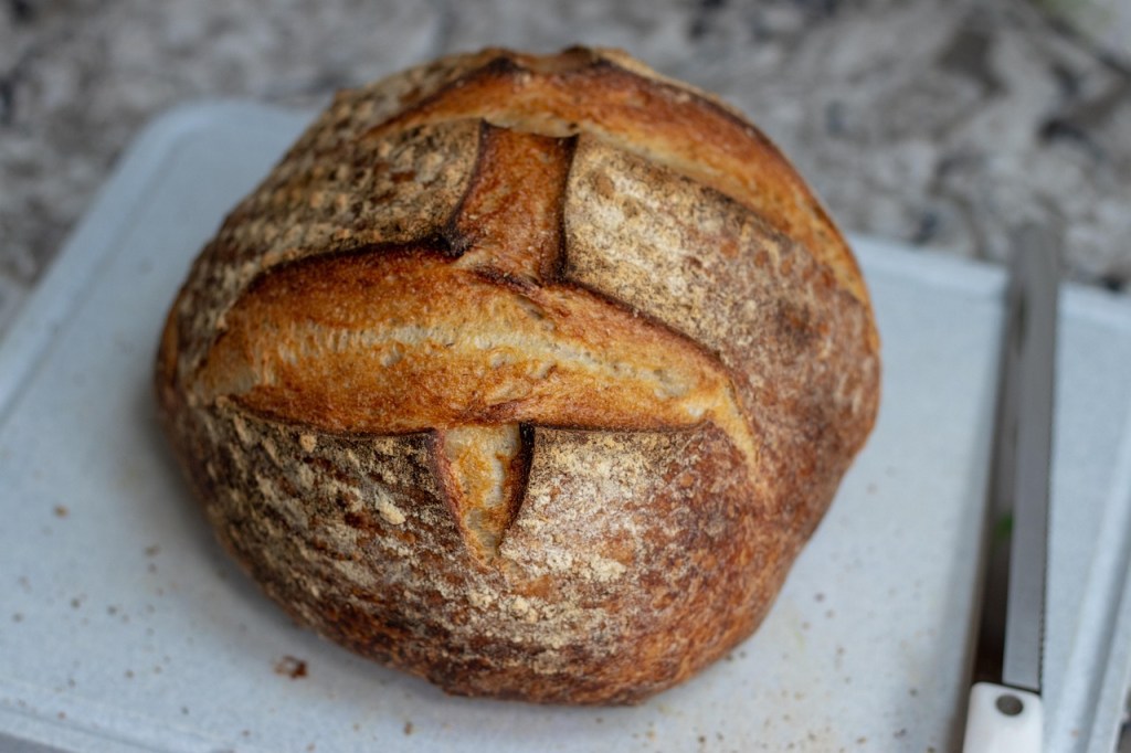 A freshly baked round loaf of sourdough bread with a golden, crusty exterior rests on a cutting board beside a bread knife.