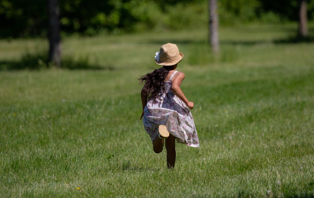 A child wearing a straw hat and a floral dress is running across a grassy field with trees in the background.