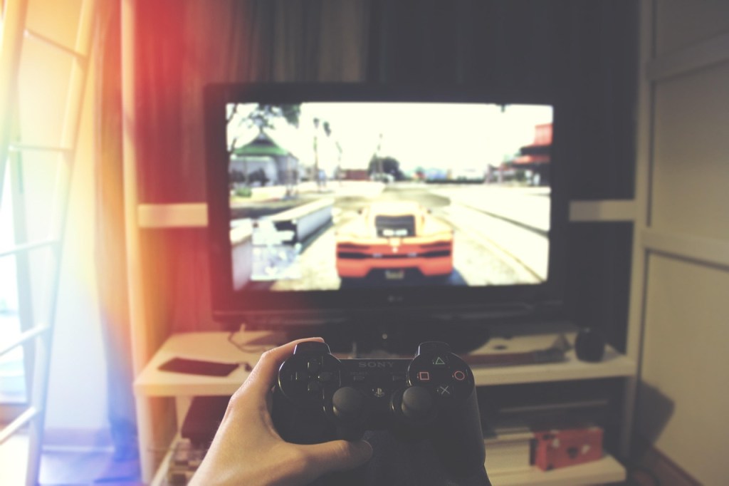 Hands holding a video game controller in front of a television screen displaying a racing game with a car on the road.