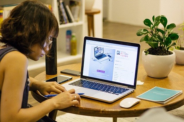 Photo is of a woman working at a table from home. She has 2 cell phones, a water bottle, calendar and a plant on her workspace. She has created a work space to eliminate distractions and create workflow.