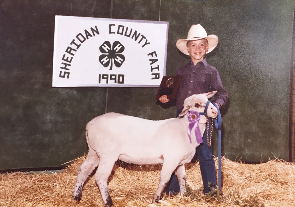 Chis Odegard stands with a sheep wearing a purple ribbon in front of a sign reading "Sheridan County Fair - 1990" with the 4-H clover emblem in the middle.