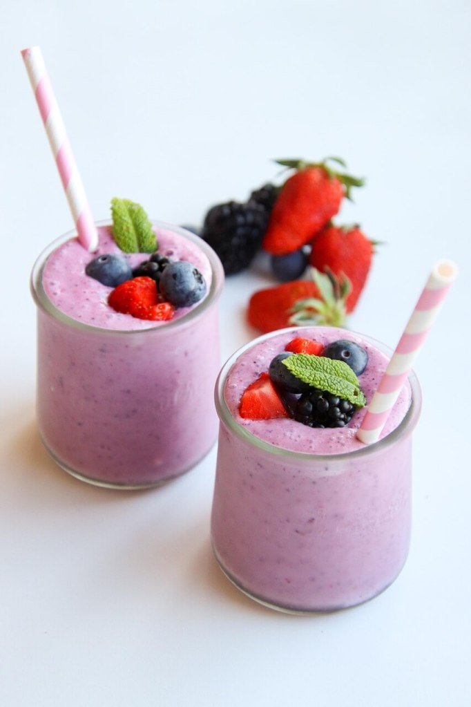 Two glasses of purple berry smoothies garnished with strawberries, blueberries, blackberries, and mint leaves, each with a striped straw, on a white surface.