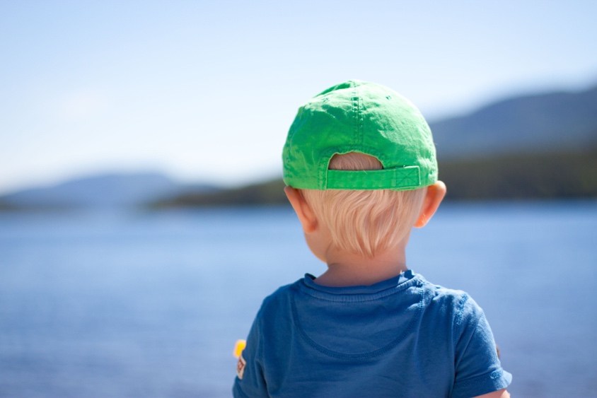 A young child wearing a green cap looks out over a lake with mountains in the background.