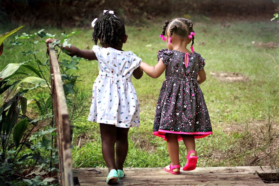 Two children walk hand in hand across a small wooden bridge in a grassy outdoor area.