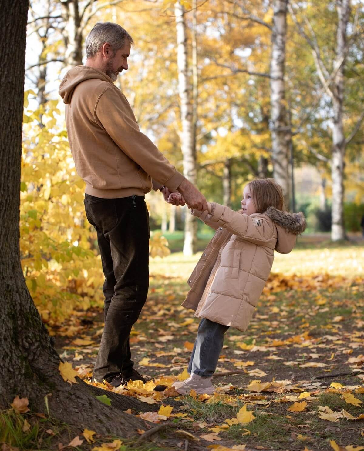 An adult and child hold hands while standing among fallen leaves in a park with autumn trees.