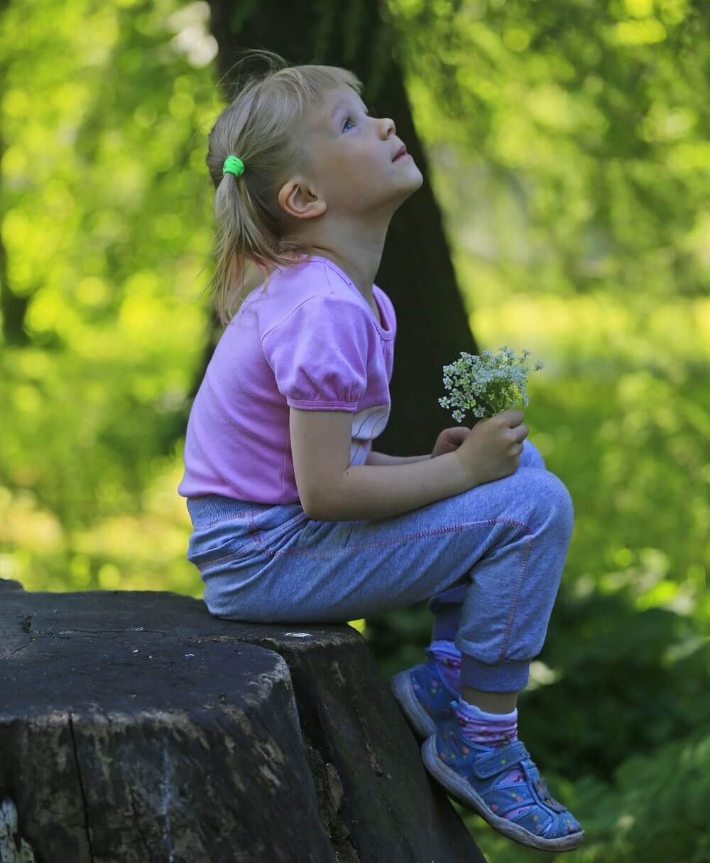 A child sits on a tree stump holding a small bouquet of flowers in a green park setting.