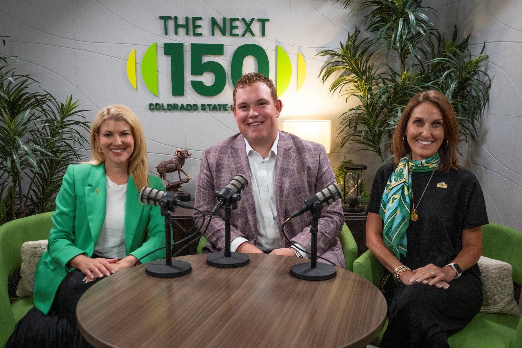 Amy Parsons, Ryer Roberts, and Jill Bramble sit around a table with microphones in a podcast-style recording setup. Behind them is a sign reading “The Next 150 Colorado State University,” with plants and decorative furnishings in the background.