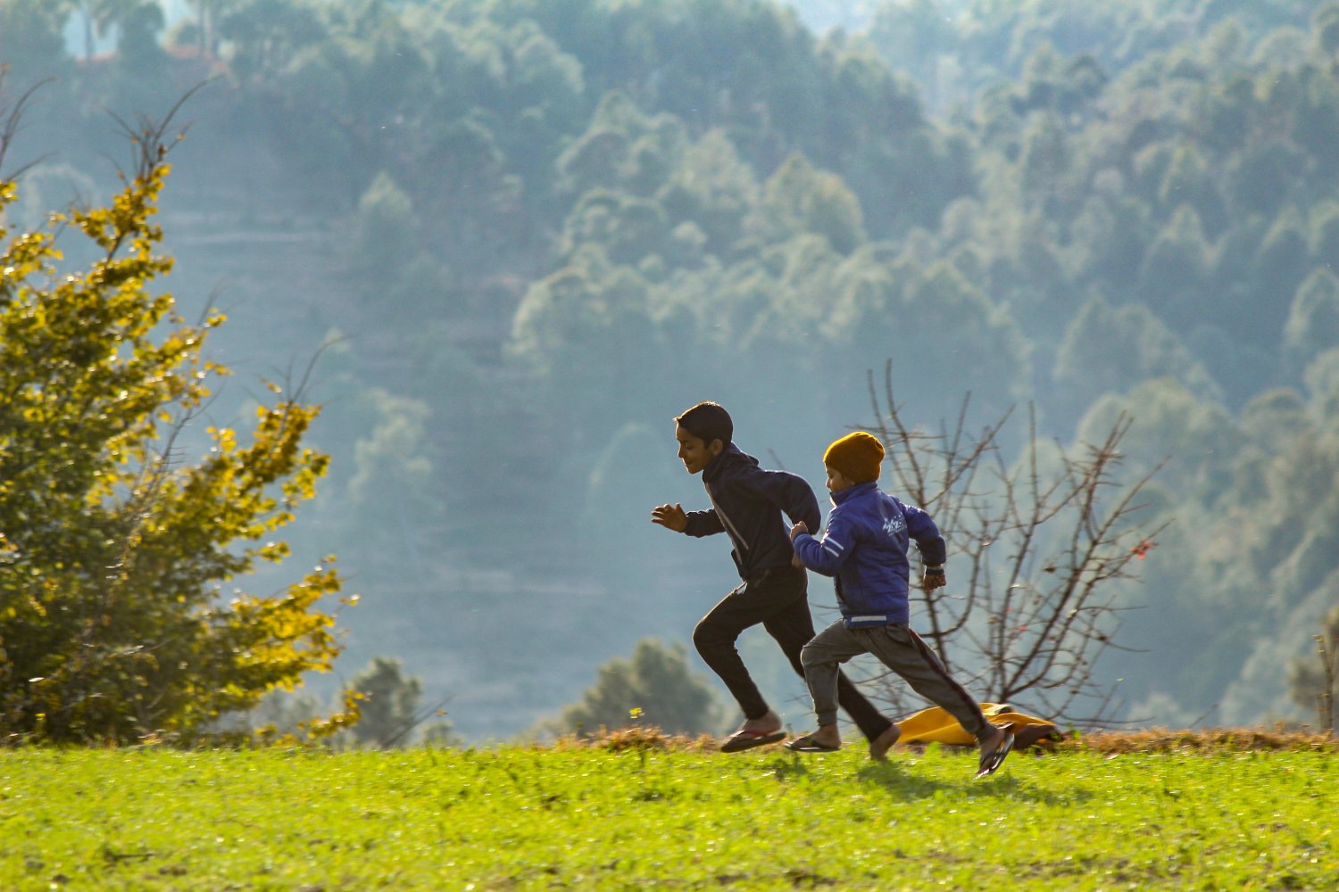 Two children run across a grassy field with trees and hills in the background.