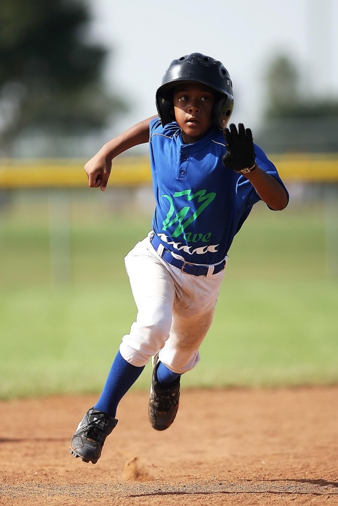 A youth baseball player wearing a helmet and uniform runs across a baseball field.